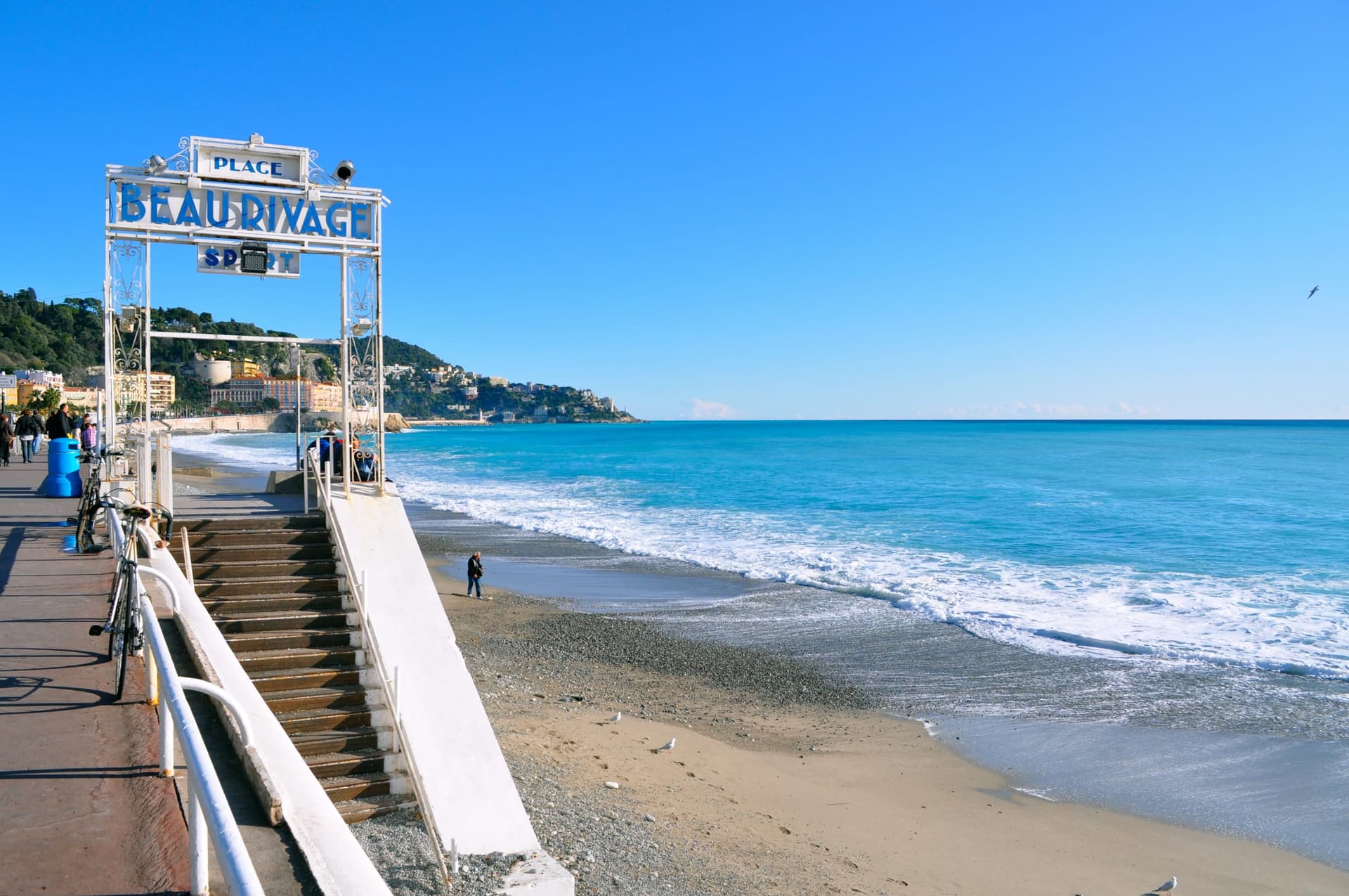 Le rivage de Nice au niveau de la plage : les galets, la mer turquoise, le portail en fer forge d'une plage de la Promenade et la colline du Chateau au fond.